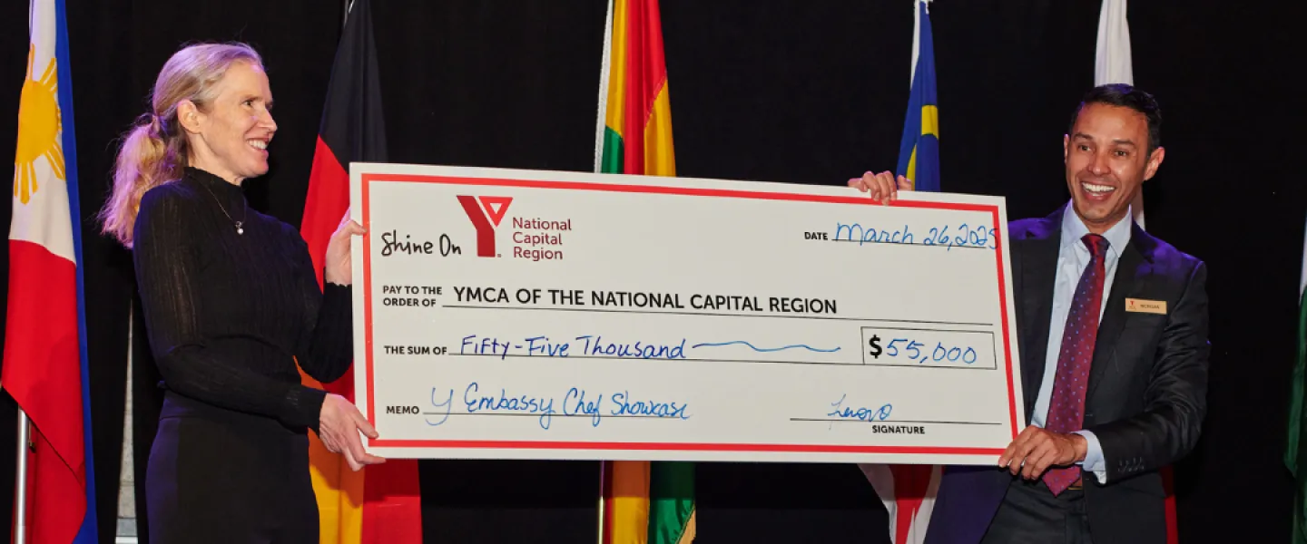 Anne Butler and Morgan Bello holding a large cheque to the YMCA of the National Capital Region for $55,000. There are several countries' flags behind them.