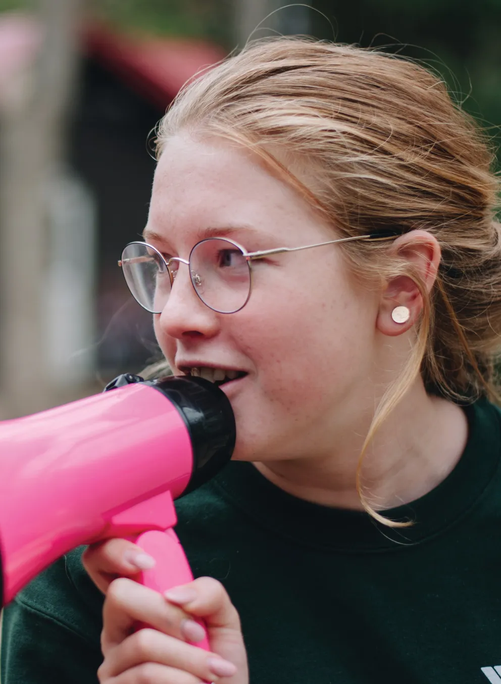 A girl with glasses enthusiastically holds a pink megaphone, ready to speak or cheer.
