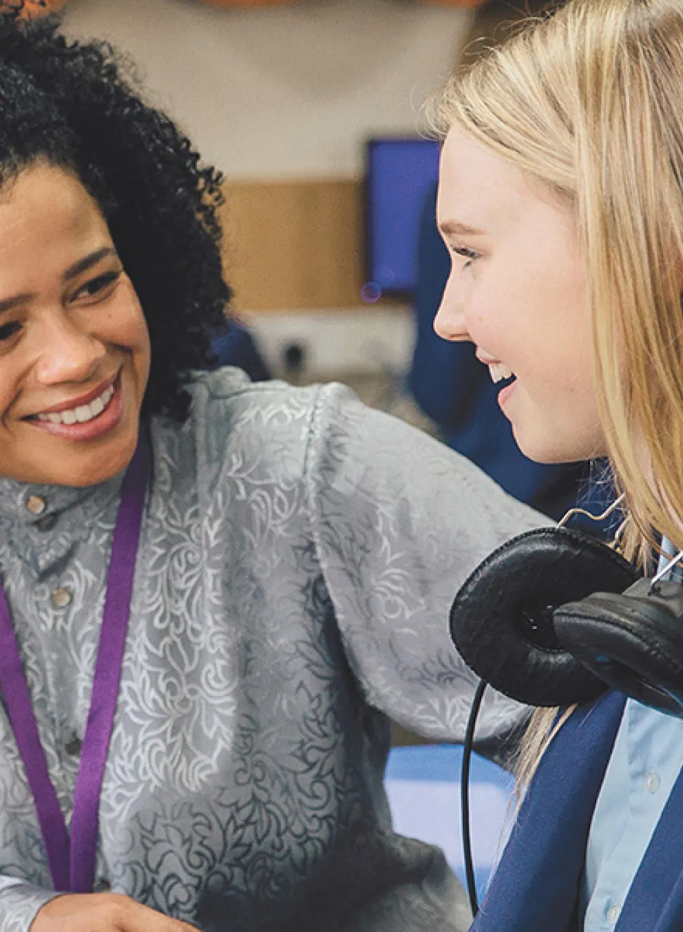 A young girl wearing headphones engages in conversation with her teacher in a classroom setting.