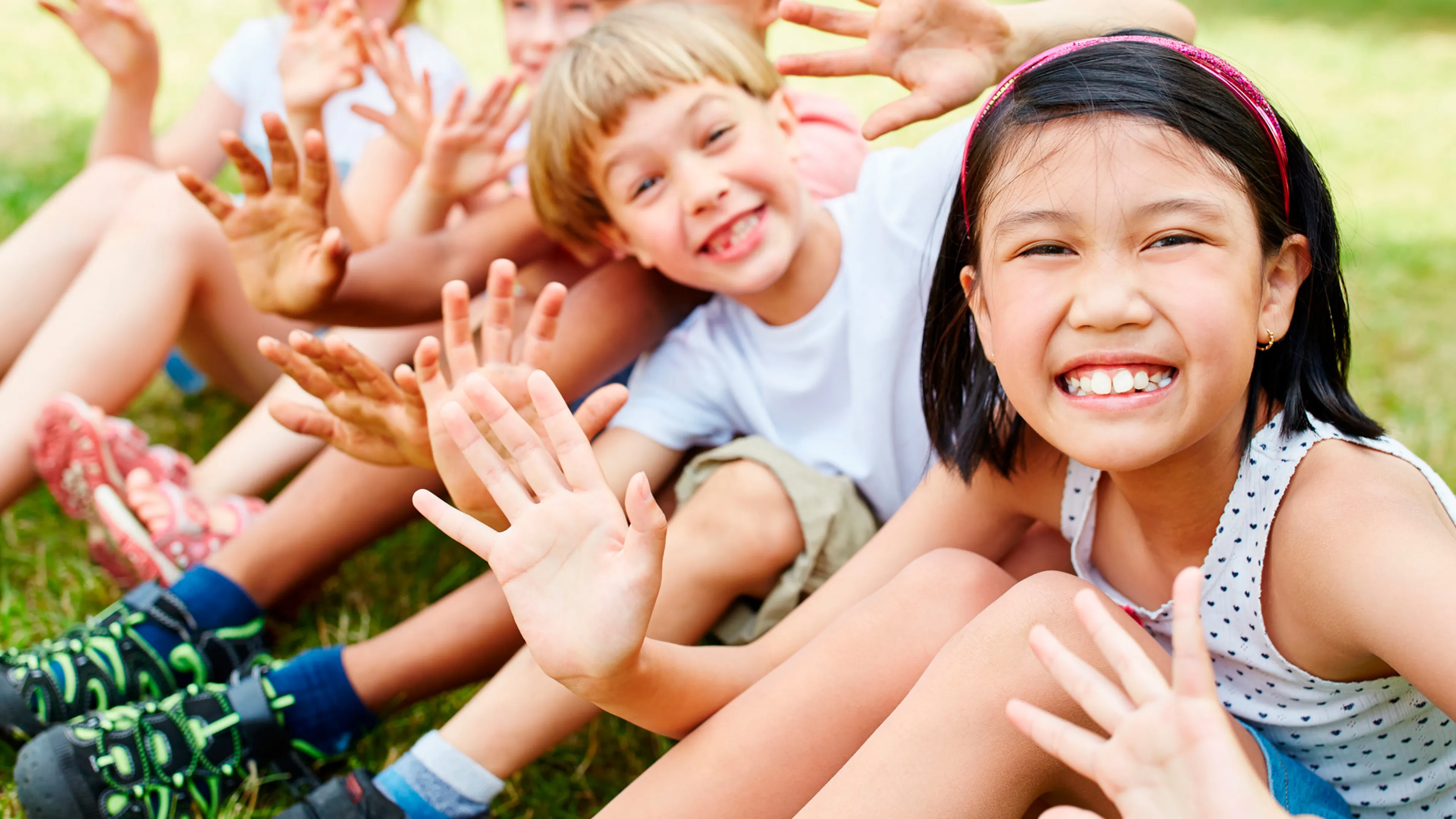 A group of children sit in a row smiling and waving.