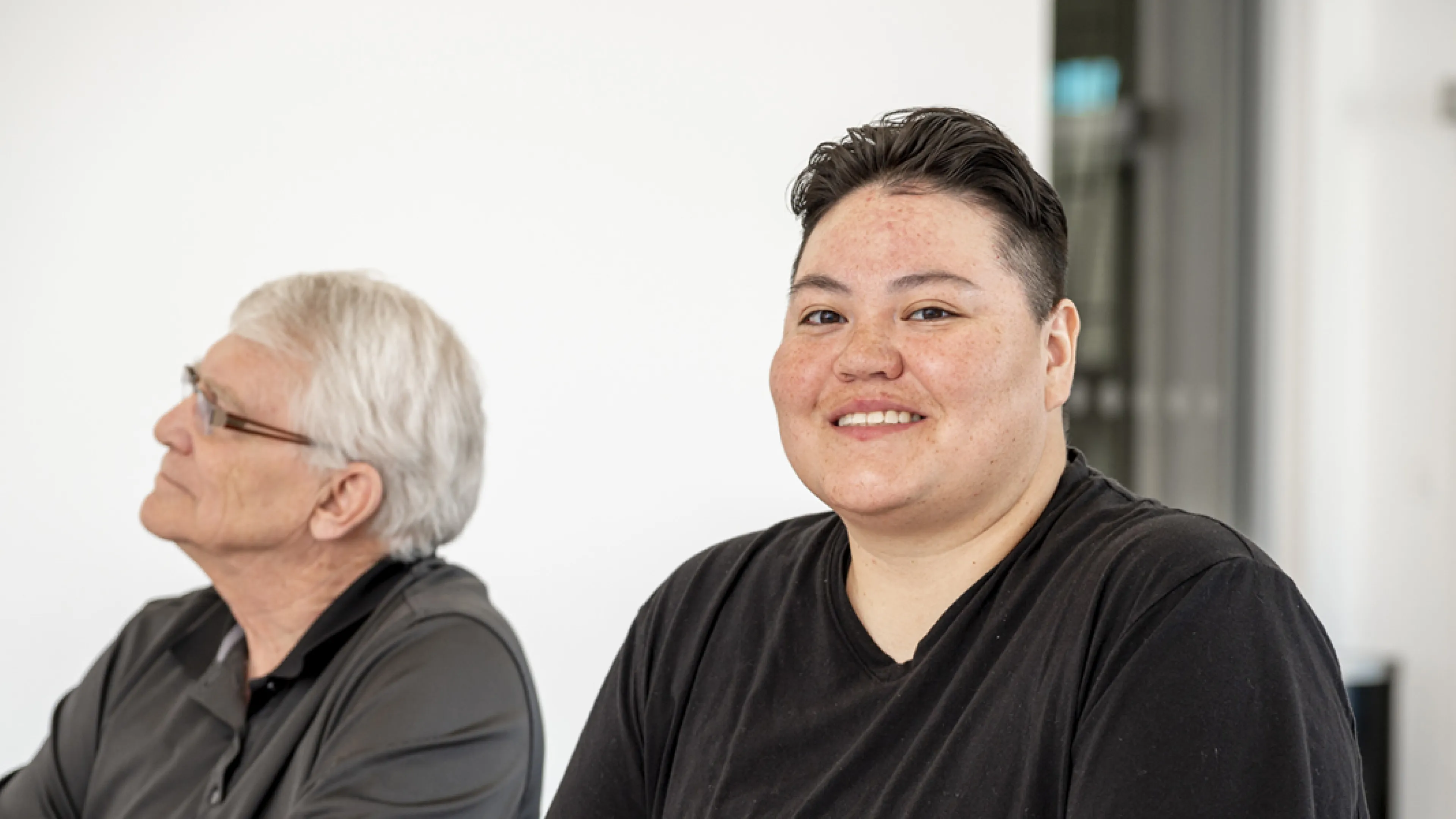 Two people sitting at a conference table. One person looks towards the presenter while the other one smiles at the camera.