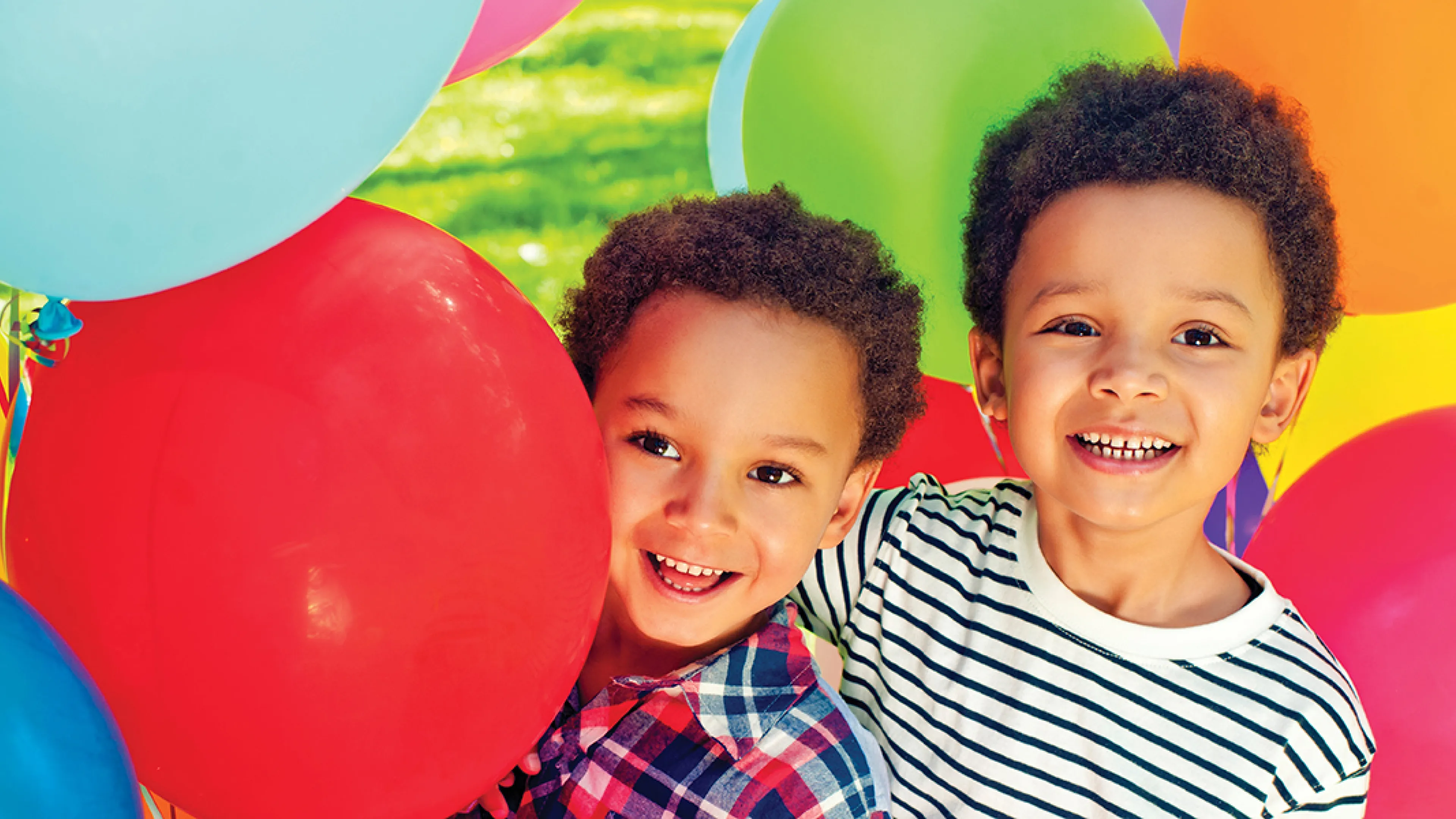 Two young boys smile joyfully in front of a vibrant display of colorful balloons.