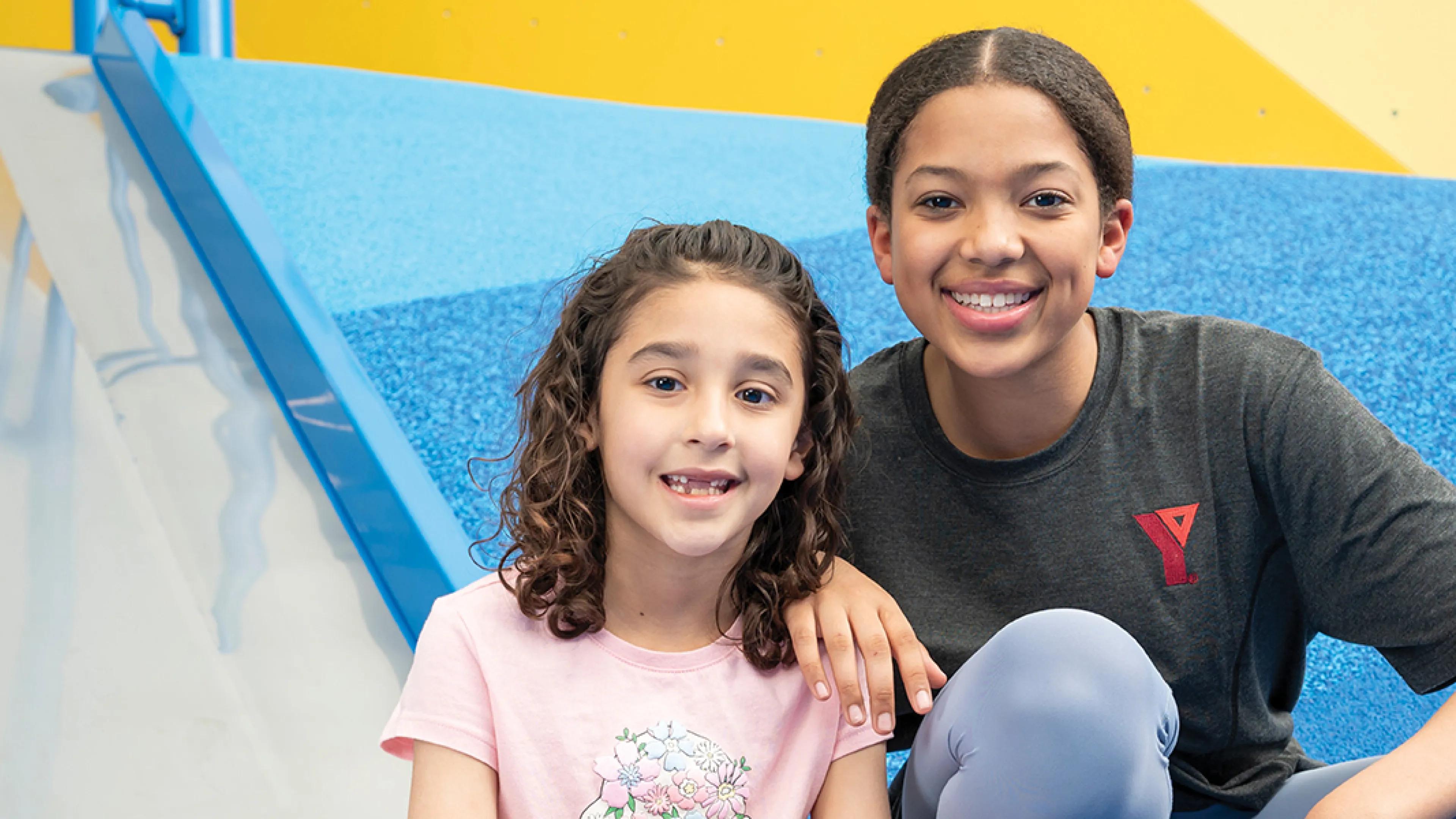 Two young girls sitting on a slide in a gym, smiling and enjoying their playtime together.