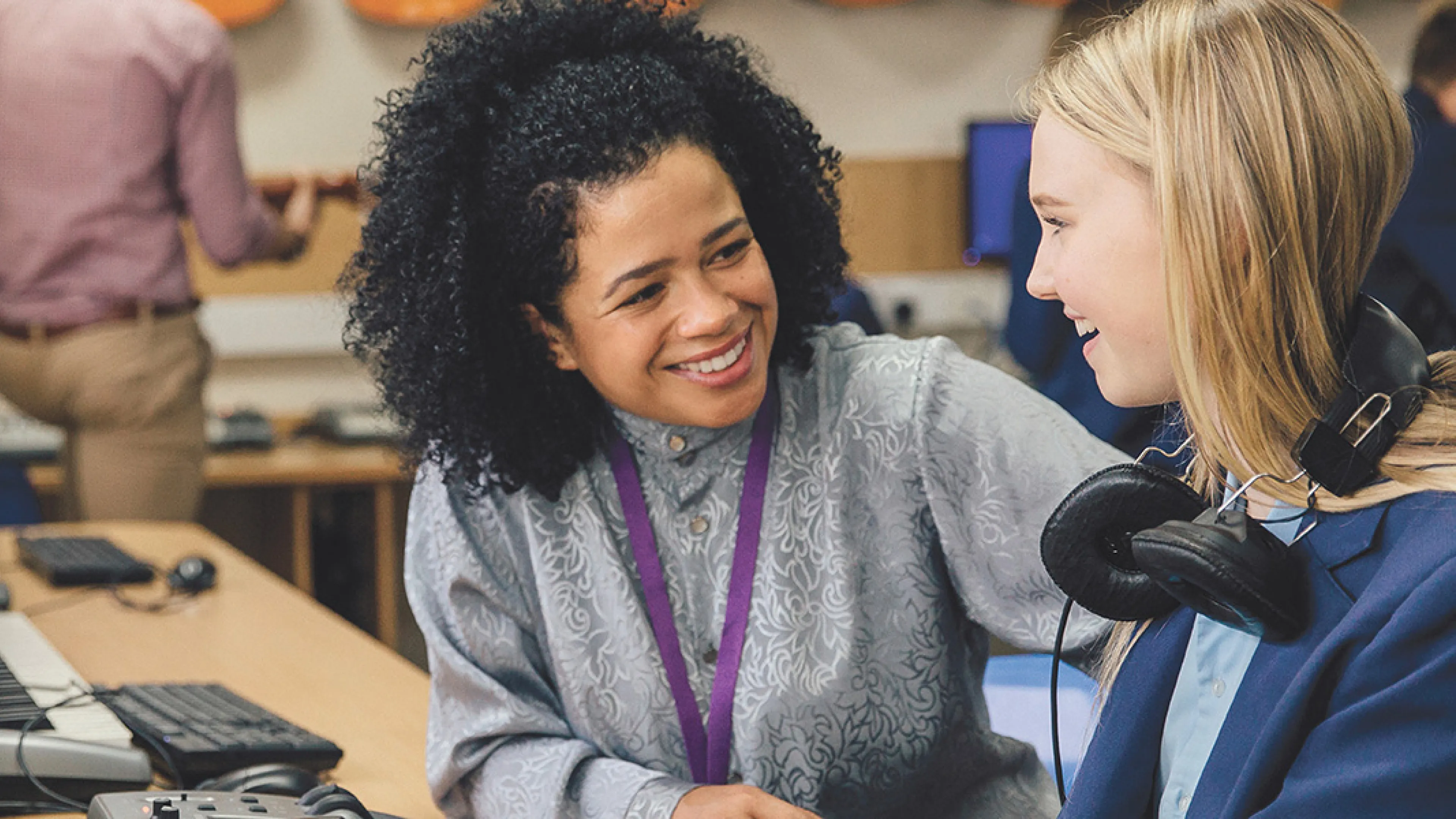 A young girl wearing headphones engages in conversation with her teacher in a classroom setting.