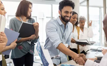 A diverse group of people in a job fair setting. A man in the foreground is smiling, shaking hands, and surrounded by others holding folders.